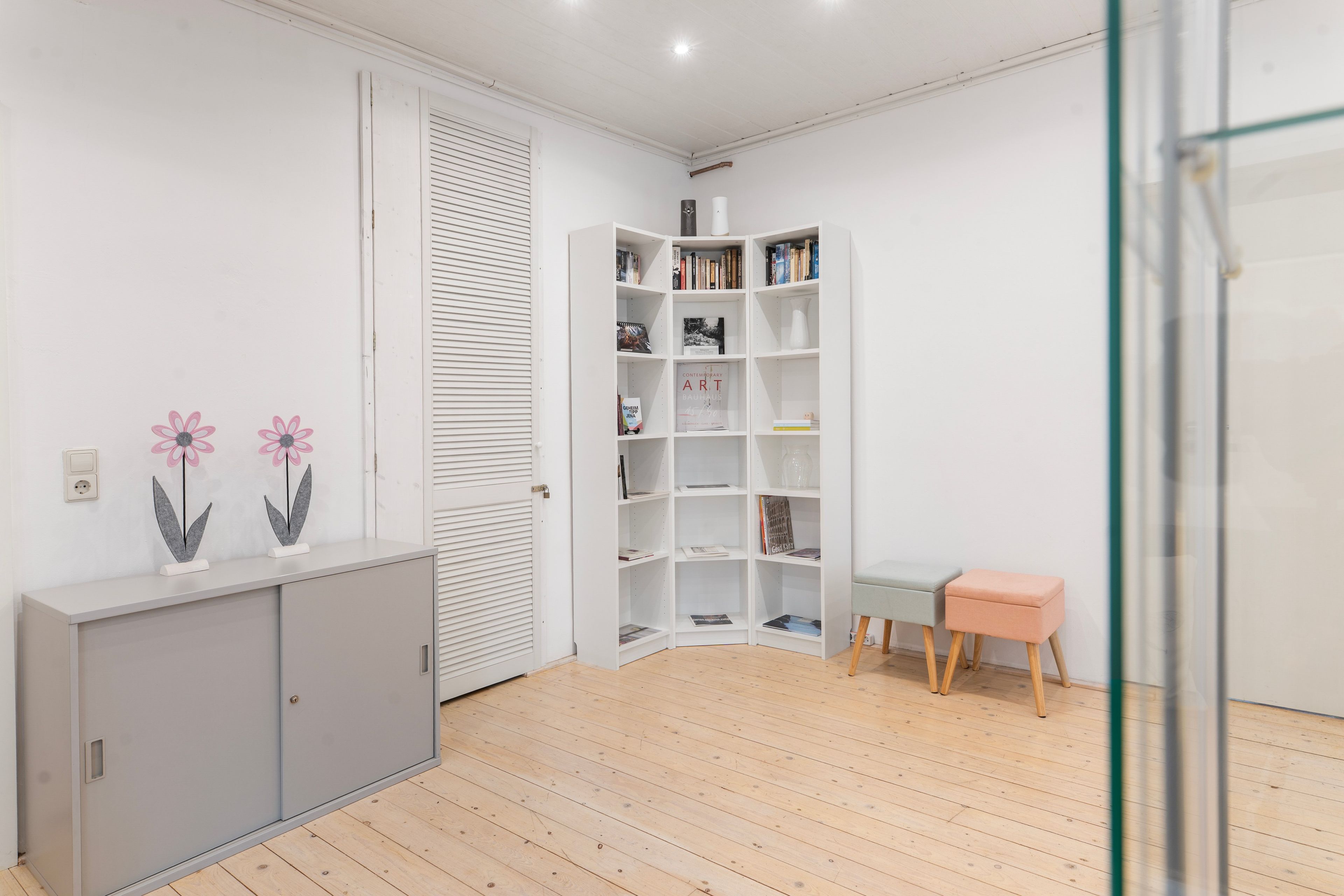 A room with a grey cabinet, a white bookshelf, a small stool, and a glass door.