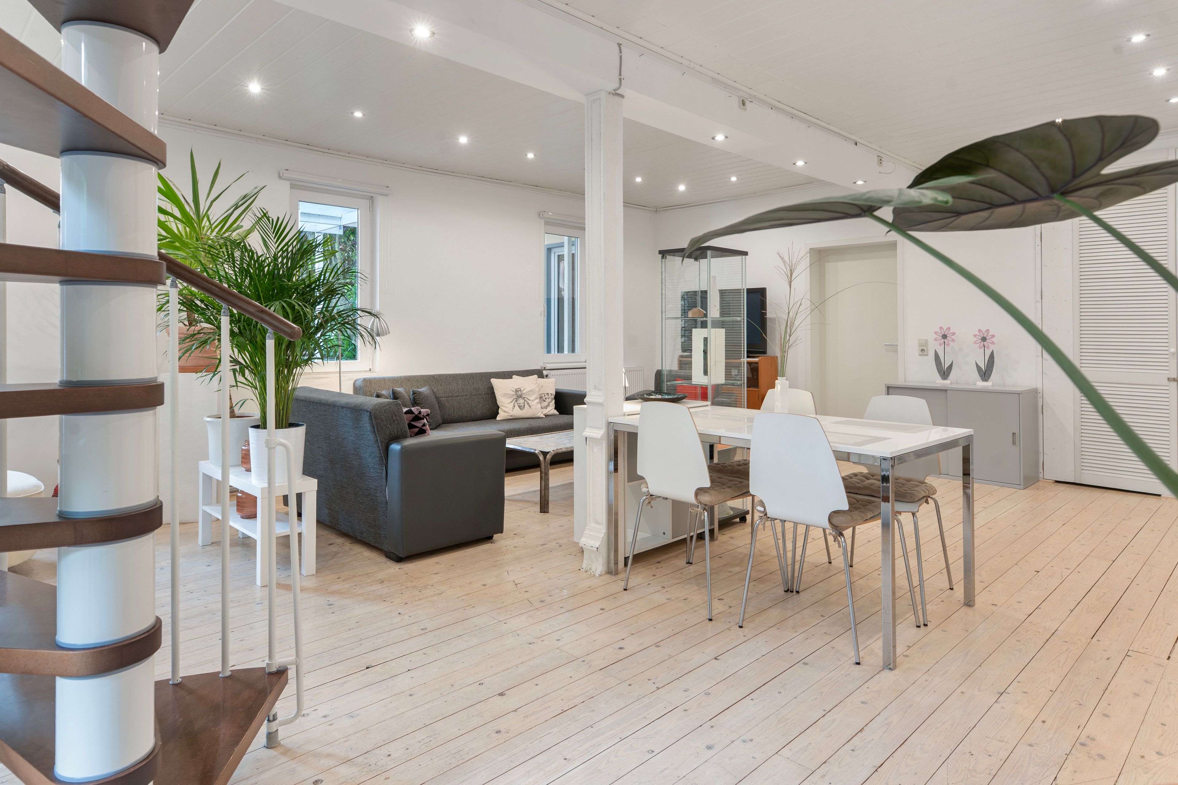 Amplia sala de estar con un sofá gris, una mesa de comedor con sillas y una planta grande en maceta.