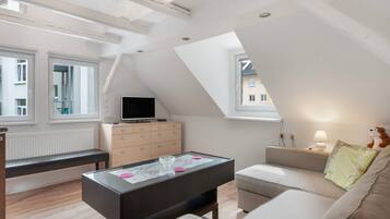 Ein modernes Wohnzimmer mit Dachstockdecke, Flachbildfernseher, Glastisch, Sofa und einer Lampe.