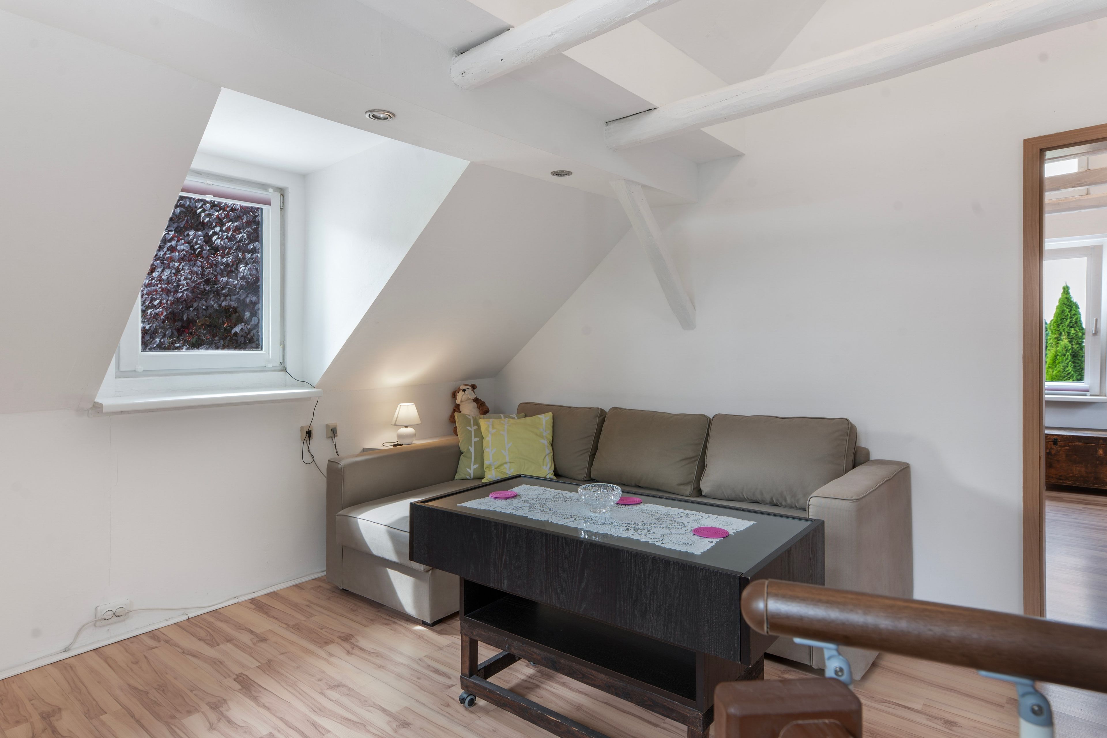Ein Zimmer mit schrägem Dach, einem Fenster mit Blick auf Bäume, einer Couch mit Kissen, einem Tisch mit Spitzenunterlage und einer Lampe.
