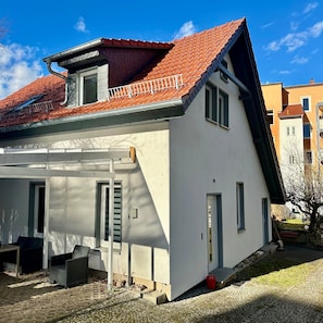 Ein eingeschossiges Haus mit einem geneigten roten Ziegeldach, einem kleinen Balkon und einem gepflasterten Auffahrt.