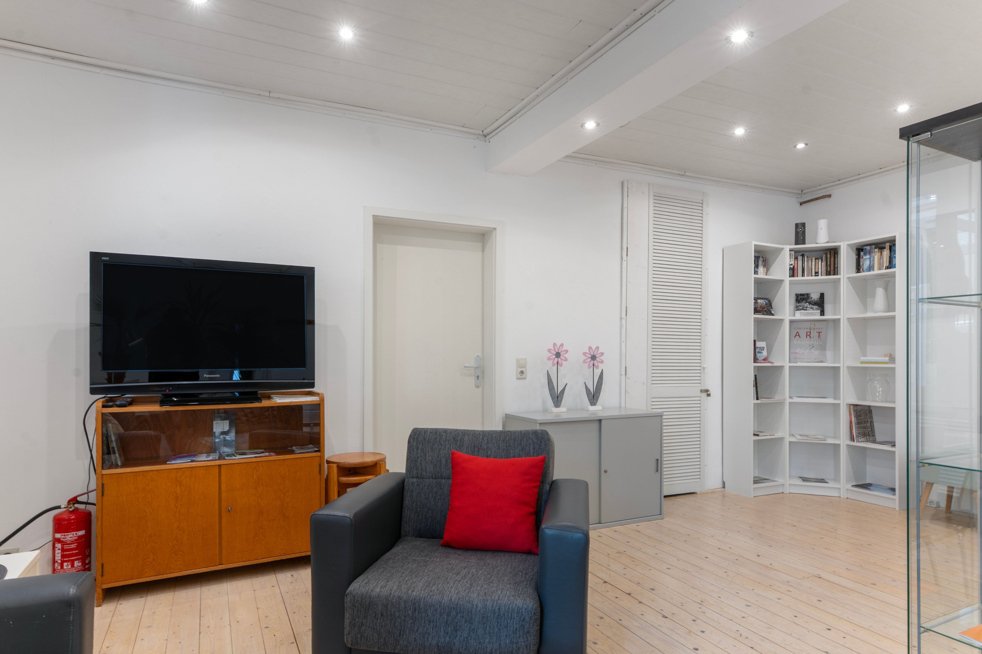 Una sala de estar moderna con un televisor de pantalla plana, un sofá gris con un cojín rojo, un mueble de entretenimiento de madera y una estantería con puertas de vidrio.