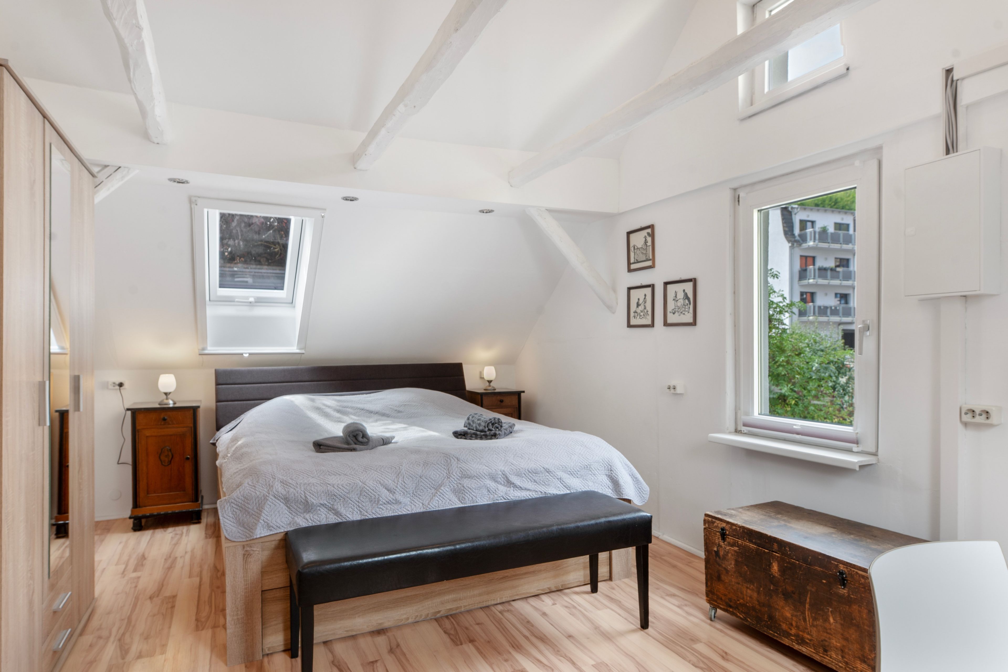 A bedroom with a bed, bench, bedside tables, a window, and a view of the outside.