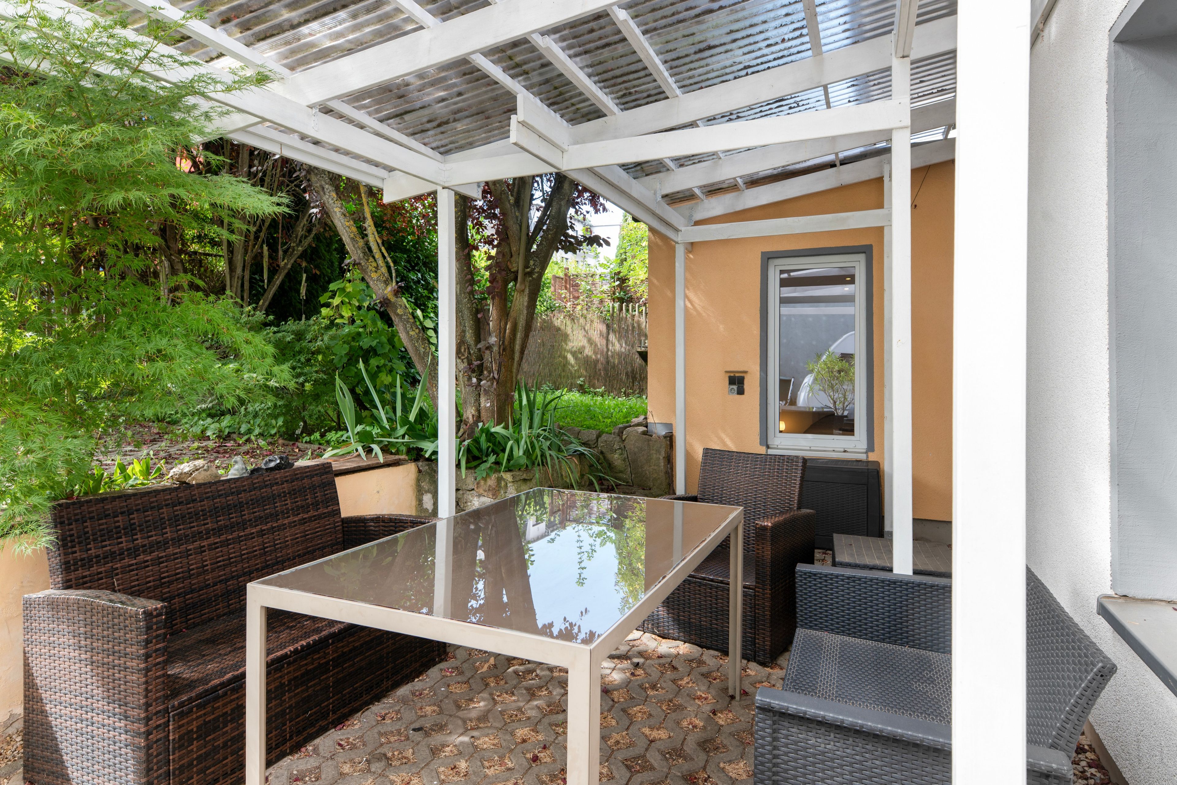 Eine Terrasse im Freien mit einem Glastisch und Korbmöbeln, umgeben von Grünpflanzen und einer Pergola.
