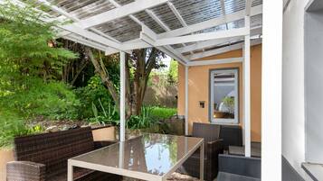 Eine Terrasse im Freien mit einem Glastisch und Korbmöbeln, umgeben von Grünpflanzen und einer Pergola.