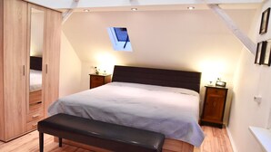 Un dormitorio con cama, una banca, armario, mesitas de noche y un ventanal pequeño.