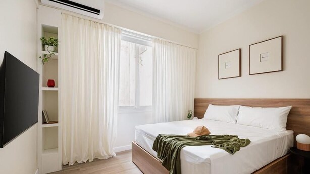 Une chambre avec un lit, une télévision, une fenêtre avec des rideaux et une étagère avec des plantes.