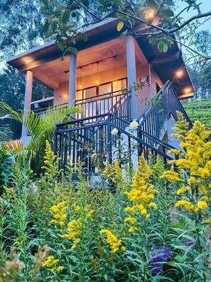 Un edificio de dos pisos con un balcón, rodeado por un jardín con flores amarillas.