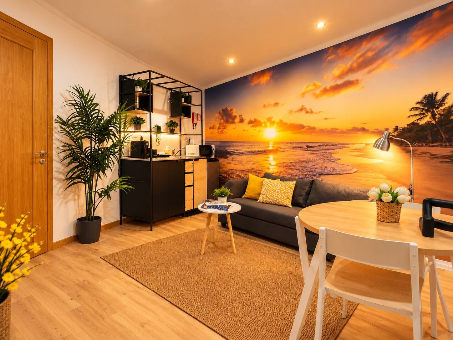 A cozy living area with a kitchenette, a sofa, a dining table, and a large sunset mural on the wall.