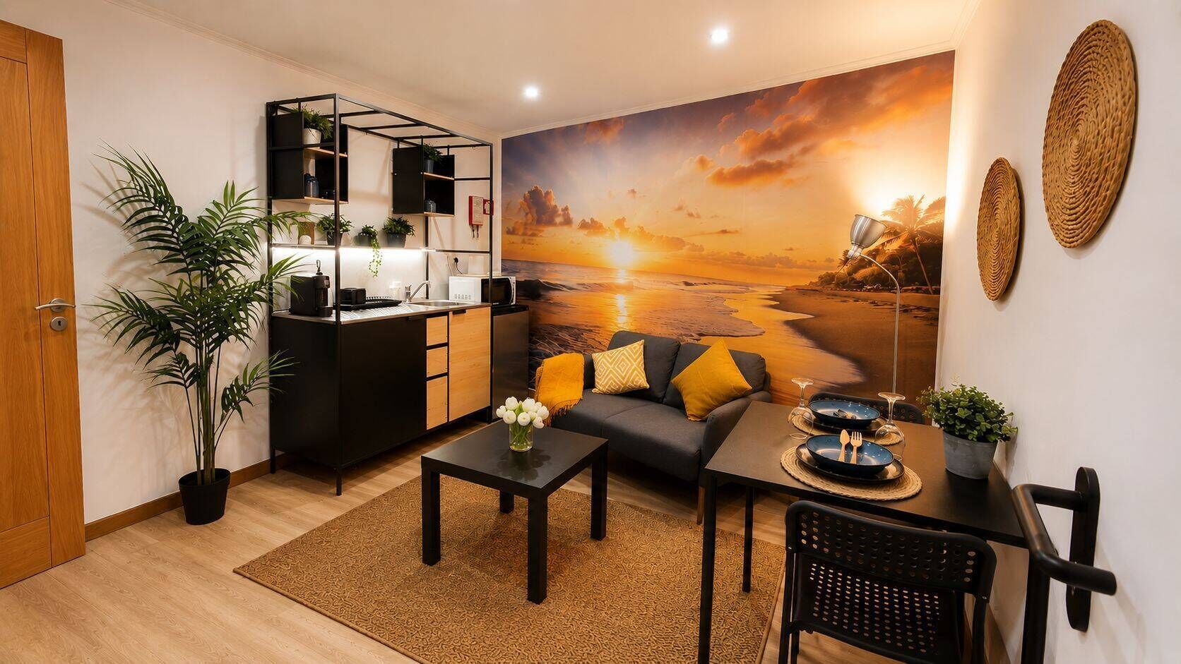 Uma sala de estar moderna com sofá, mesa de centro e área de refeições. Há uma cozinha com bancada preta e prateleiras. Uma grande pintura de um pôr do sol sobre o oceano decora a parede.
