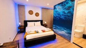 Ein modernes Hotelzimmer mit einem großen Bett, einem Nachttisch, einem an der Wand montierten Fernseher und einem durchgehenden Türbogen einsehbaren Badezimmer.