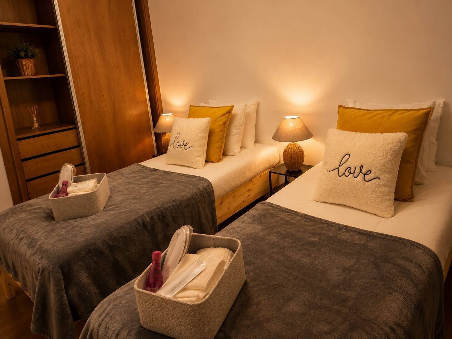 Une chambre d’hôtel avec deux lits, chacun agrémenté d’un oreiller sur lequel est brodé le mot « amour ». Il y a des tables de chevet avec des lampes et une petite étagère avec une plante.