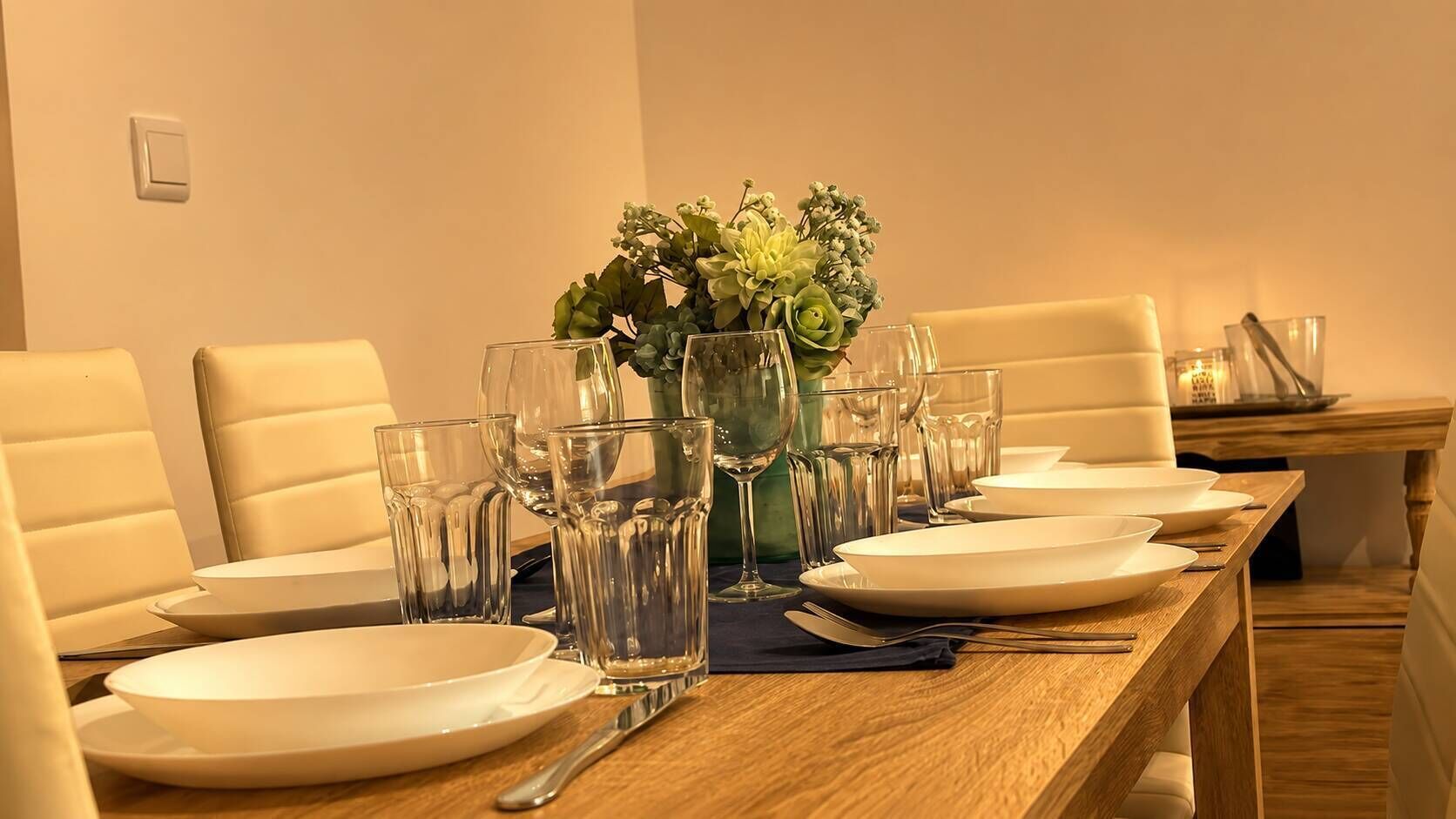 Una mesa de comedor lista con platos, vasos y cubiertos, con un florero de plantas verdes en el centro.