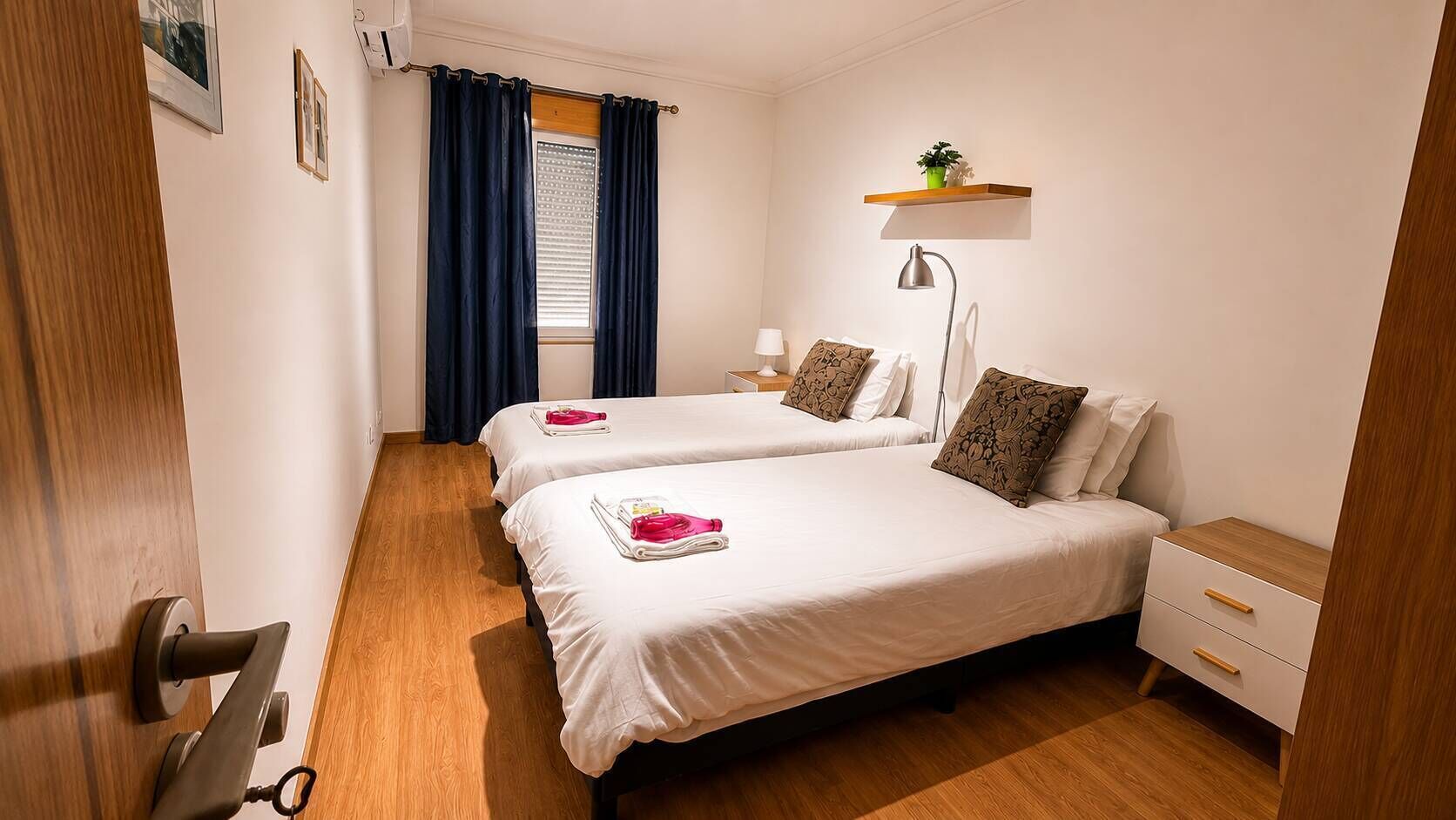 Habitación de hotel con dos camas, una mesita de noche, una lámpara, un estante con una planta y una ventana con cortinas.
