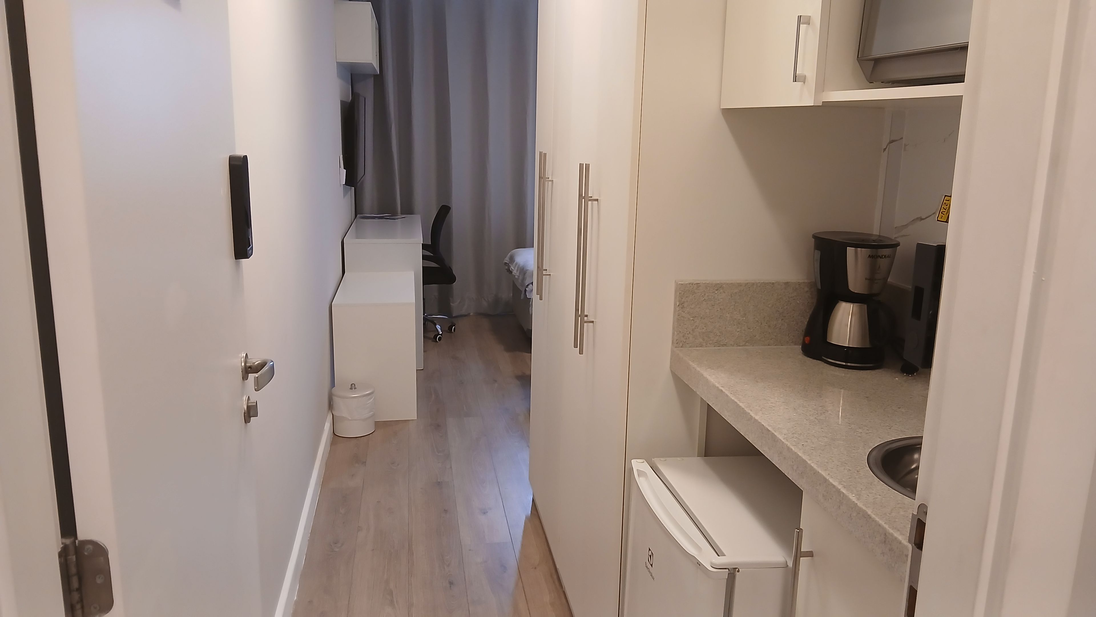 Um corredor estreito leva a um quarto com uma cama, uma escrivaninha e uma cadeira. Uma pequena cozinha compacta com cafeteira e lixeira é visível.