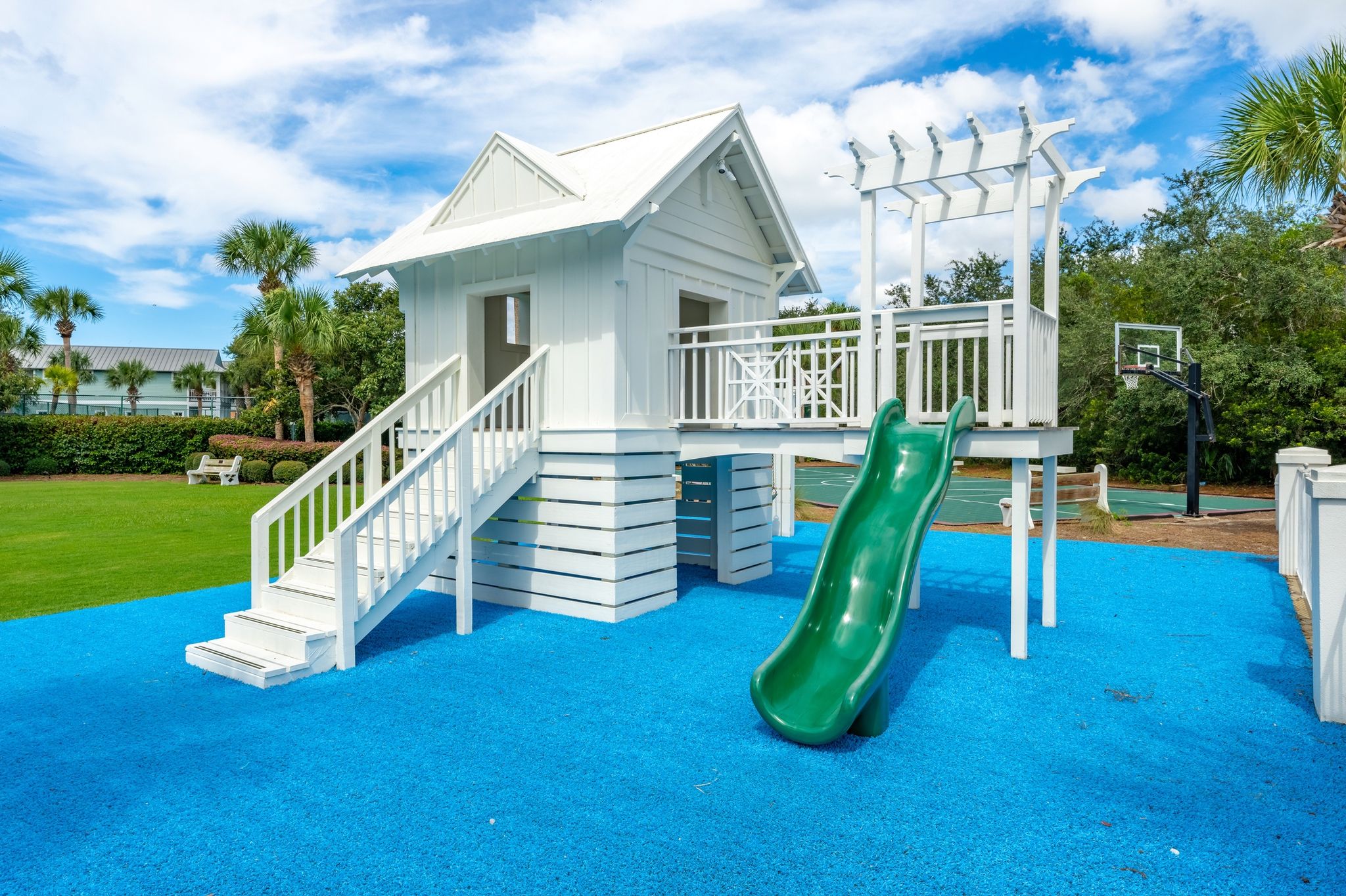Un parque infantil con un tobogán, una pequeña construcción en forma de casa blanca y un aro de baloncesto.