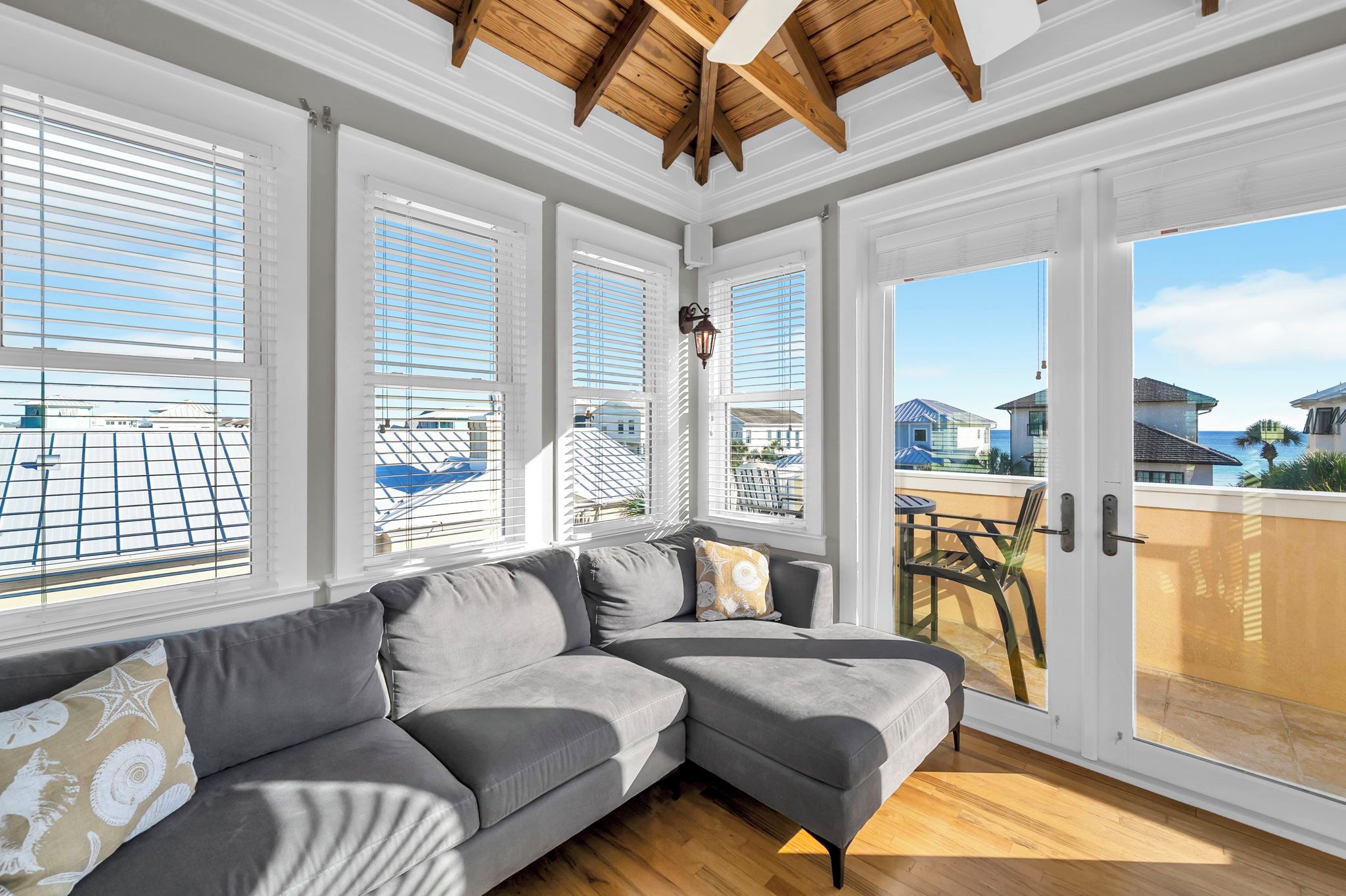 A room with a sectional sofa, large windows with blinds, a wooden ceiling, and a view of the ocean.