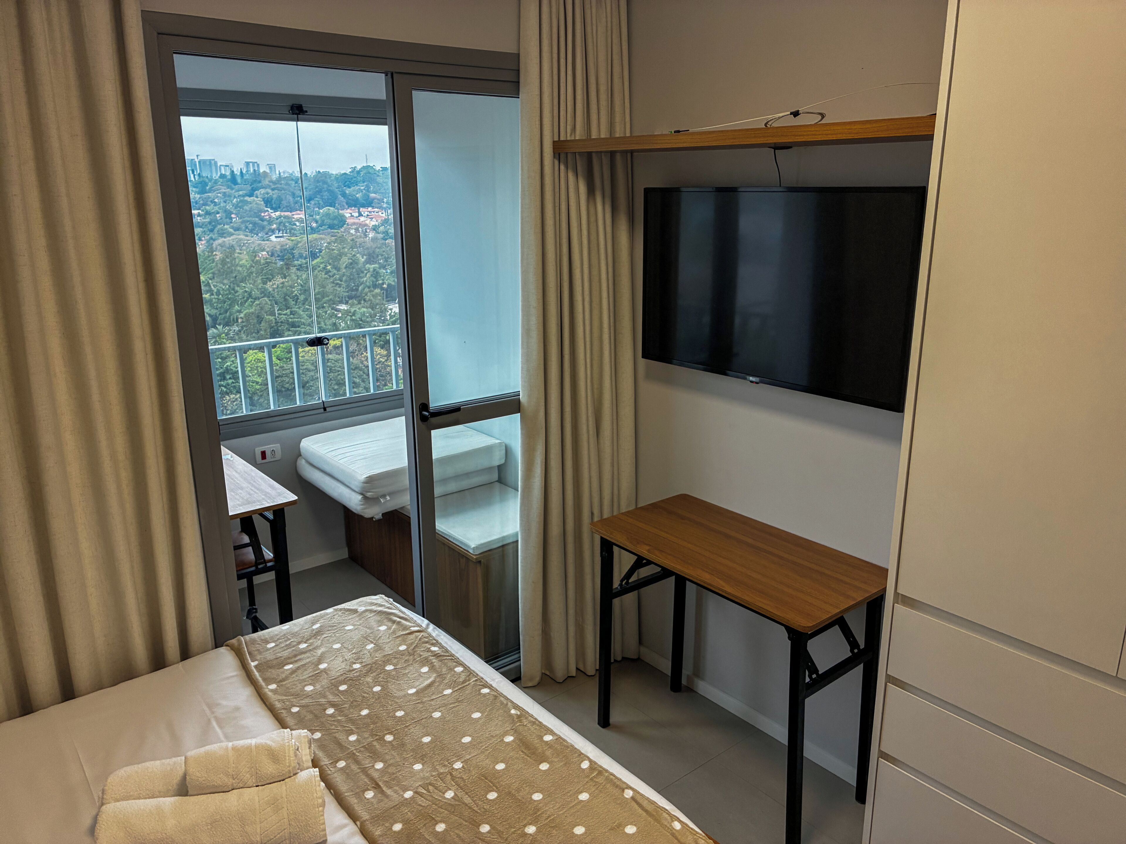 Une chambre d’hôtel comprenant un lit, un bureau, une télévision, un balcon avec vue sur la ville et une fenêtre avec des rideaux.