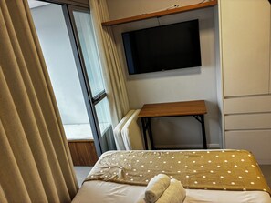 Ein Hotelzimmer mit einem Bett, einem an der Wand befestigten Fernseher, einem kleinen Holztisch und einem Fenster mit Vorhängen.