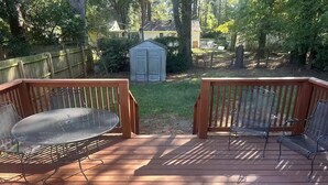 Eine Holzterrasse mit Metallstühlen und einem Tisch, umgeben von einem Holzzaun und Bäumen.