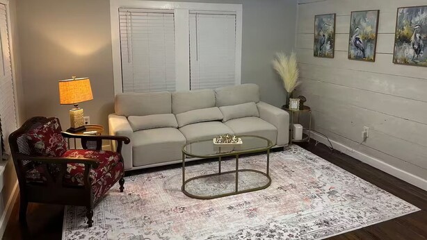 Ein Wohnzimmer mit Sofa, Couchtisch, Lampe, Sessel, Teppich und Wandbildern.