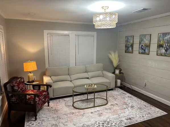A living room with a sofa, a coffee table, a lamp, a chair, a rug, and wall art. - Pixie’s Haven A Home Away From Home (Monroe)