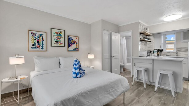 Moderni hotellihuone, jossa on sänky, yöpöytävalaisimet, kylpyhuone, pieni keittiönurkkaus ja taideteoksia seinällä.