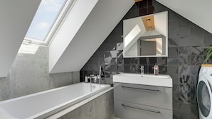 Ein modernes Badezimmer mit Oberlicht, einer freistehenden Badewanne, einem Waschtisch mit Spiegel und einer Waschmaschine.