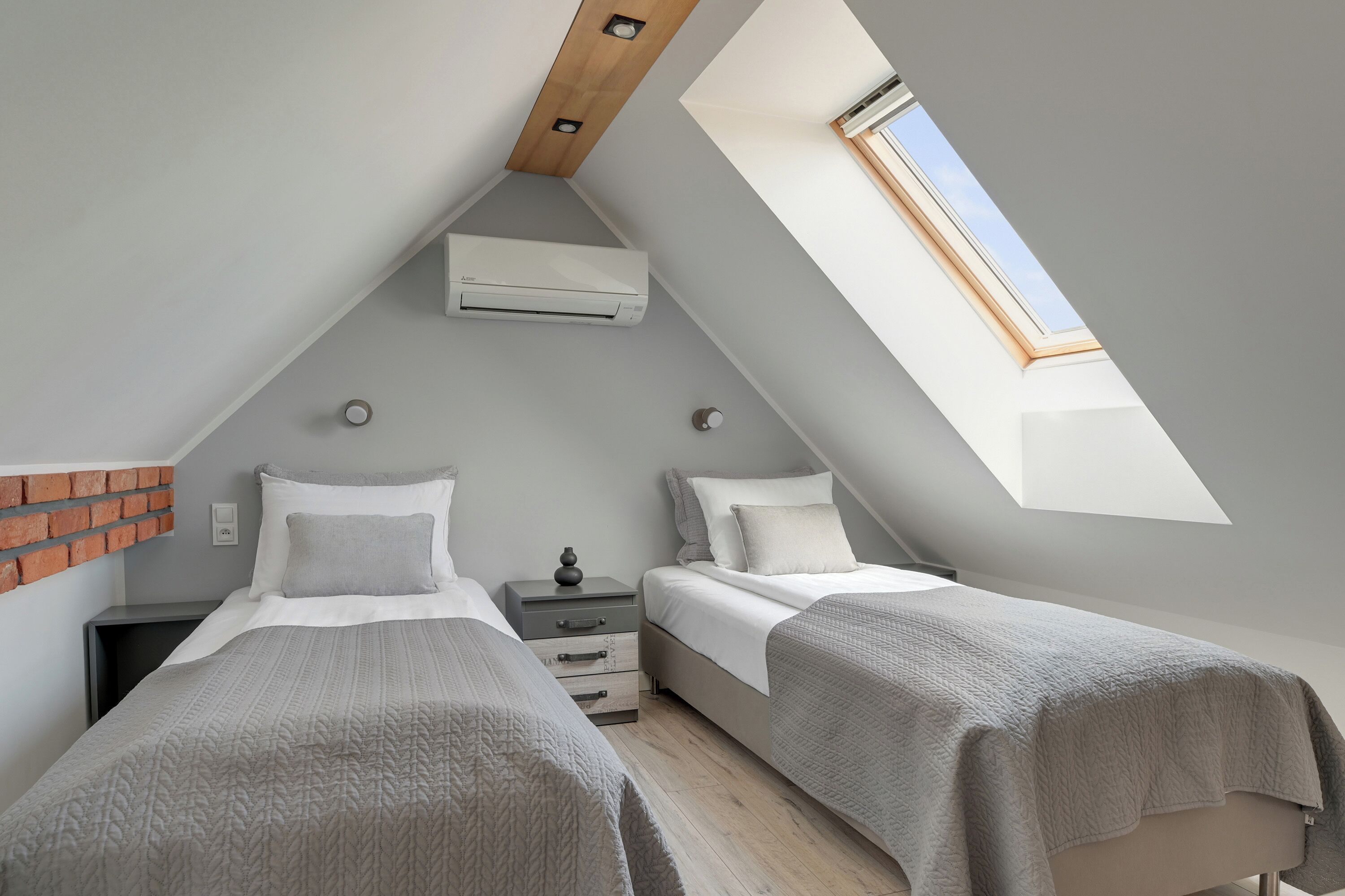 A bedroom with two beds, a skylight, an air conditioning unit, and a brick accent wall.