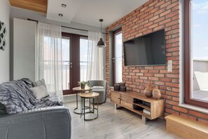 A modern living room with a brick wall, a flat-screen TV, a grey sofa, a small round table, and a window with curtains. - Waterlane Island - Pool Sauna and Gym (Gdańsk)