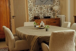 Un comedor con una mesa dispuesta para cuatro personas, rodeada de sillas. Sobre la mesa hay un florero decorativo con una planta y una pequeña figurita de un pájaro. La habitación tiene una puerta de madera, un radiador y un papel tapiz estampado.