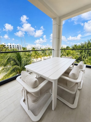 Un balcon avec une table blanche et des bancs, d’où l’on peut admirer un paysage urbain avec des immeubles et des palmiers.