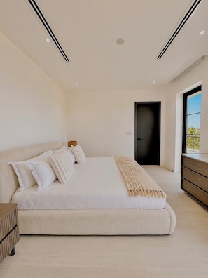 Une chambre à coucher moderne, avec un grand lit, une table de chevet, une commode et une porte donnant sur une autre pièce.