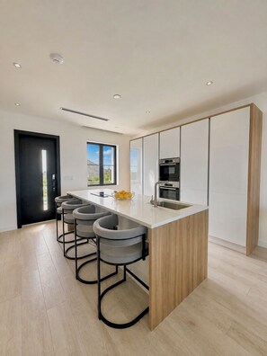 A modern kitchen with a central island, bar stools, a built-in oven, and a window with a view. - The Junkanoo Condo (Nassau)