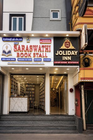 A book stall and a hotel named 'Saraswathi Book Stall' and 'Joliday Inn' with a signboard indicating amenities like CBSE & NCERT books, Tamil Nadu text books, notebooks, stationery, and luxury rooms. - RMA Joliday Inn (Puducherry)