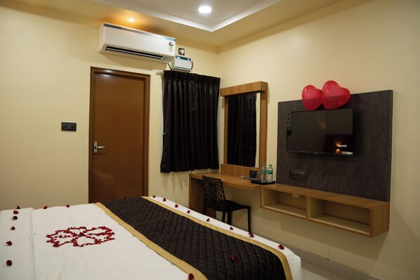 A hotel room with a bed, a desk with a chair, a flat-screen TV, an air conditioning unit, and a door.