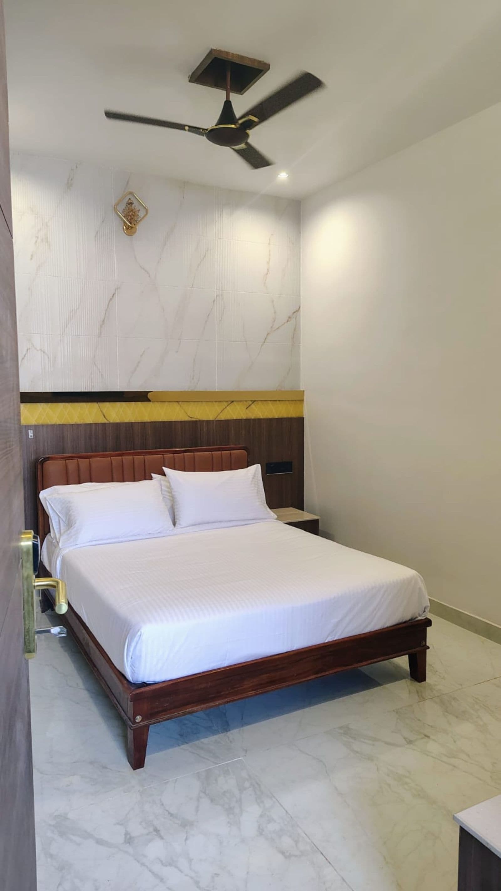 Una habitación de hotel con una cama, una mesita de noche, una lámpara de pared y un ventilador de techo.