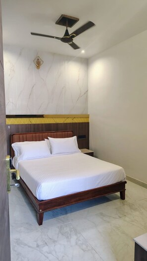 A hotel room with a bed, a nightstand, a wall-mounted light fixture, and a ceiling fan. - MKT Shanthinivas (Rameshwaram)