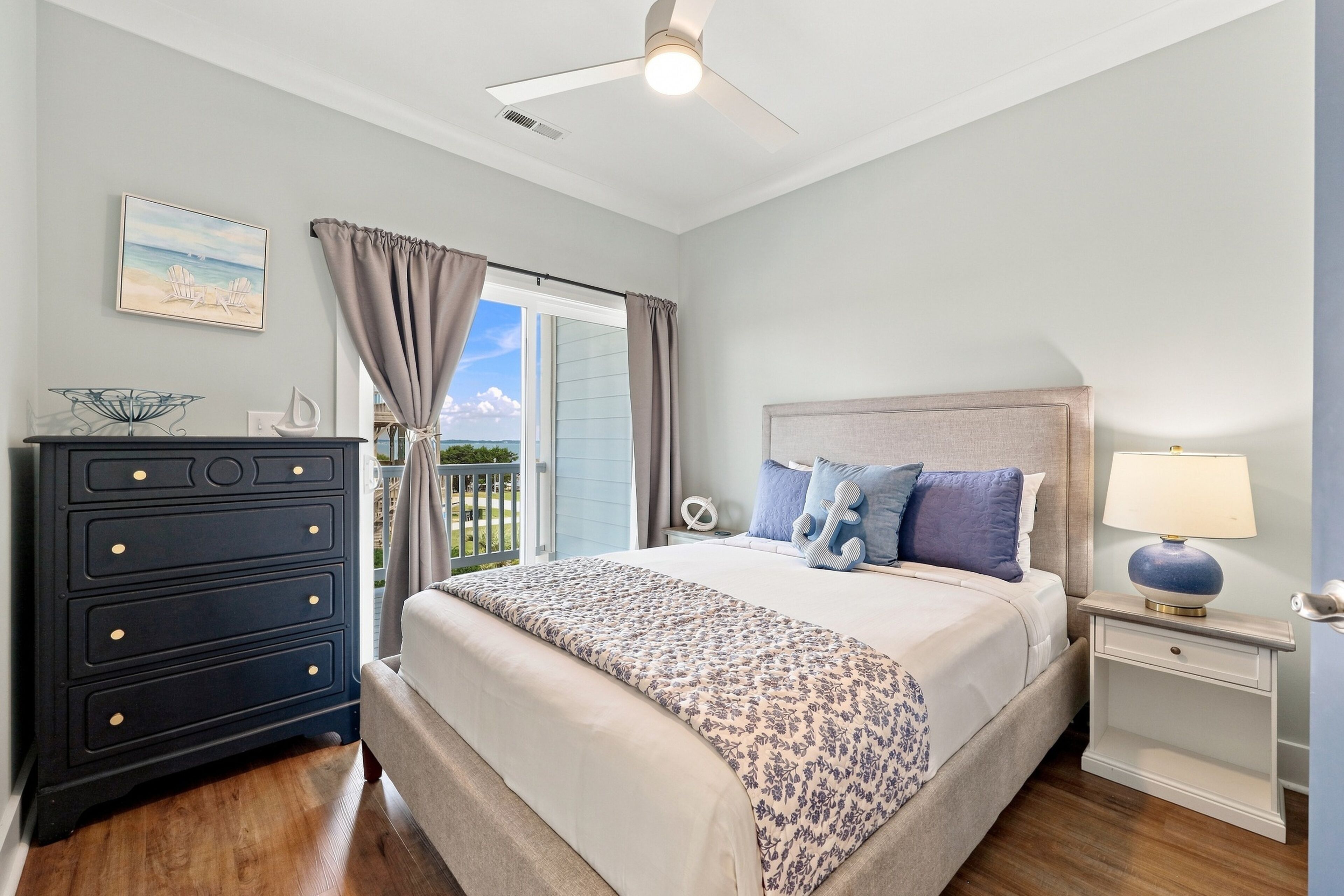 A bedroom with a bed, a dresser, a nightstand, a lamp, and a view of the outdoors.
