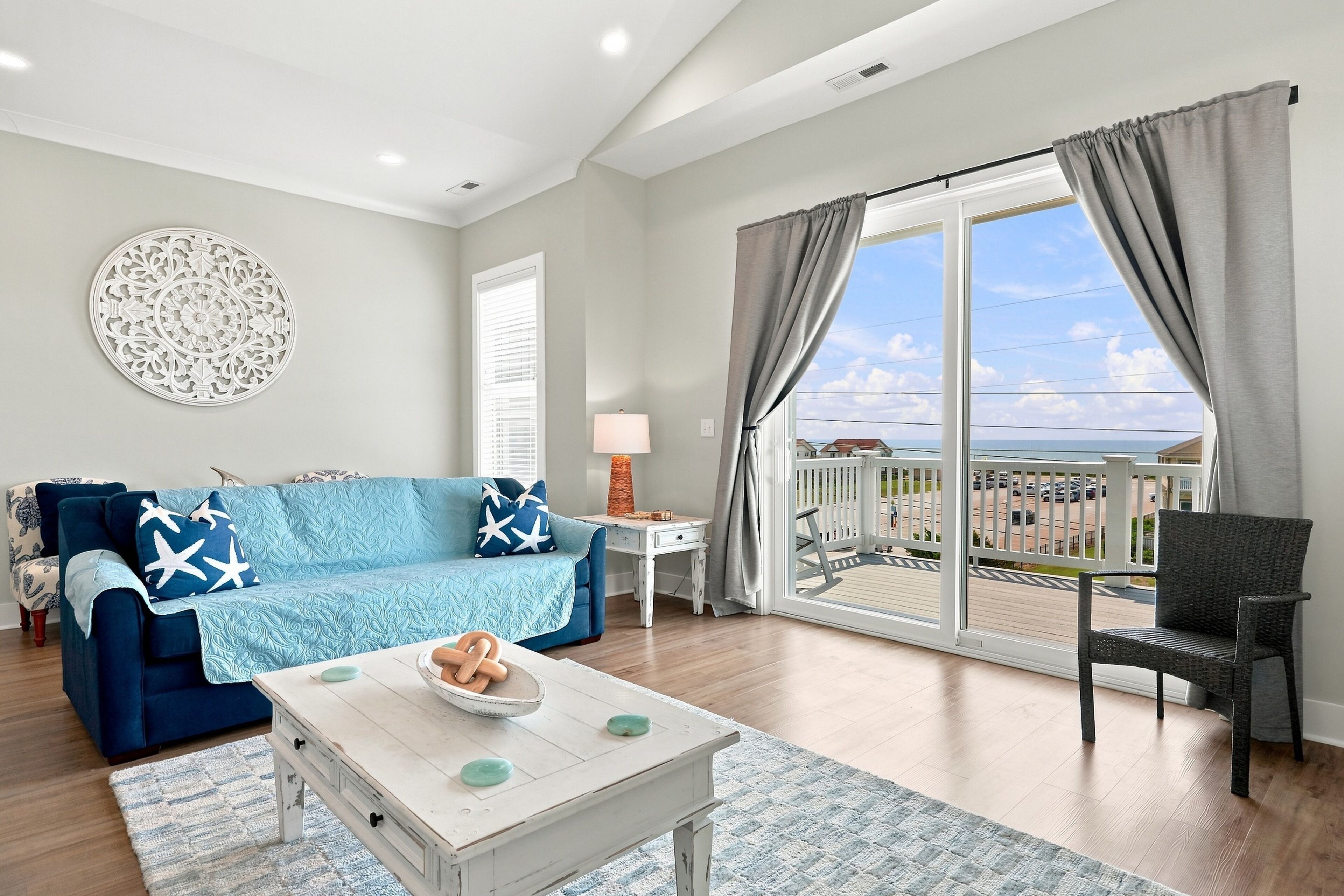 A living room with a blue sofa, a coffee table with decorative items, a white wall with a circular design, and a balcony with a view.