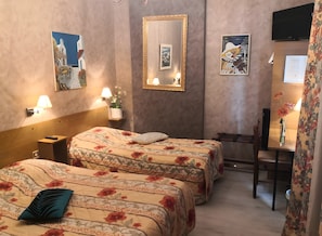 A hotel room with two beds, a desk, a chair, a mirror, wall-mounted lamps, and artwork.