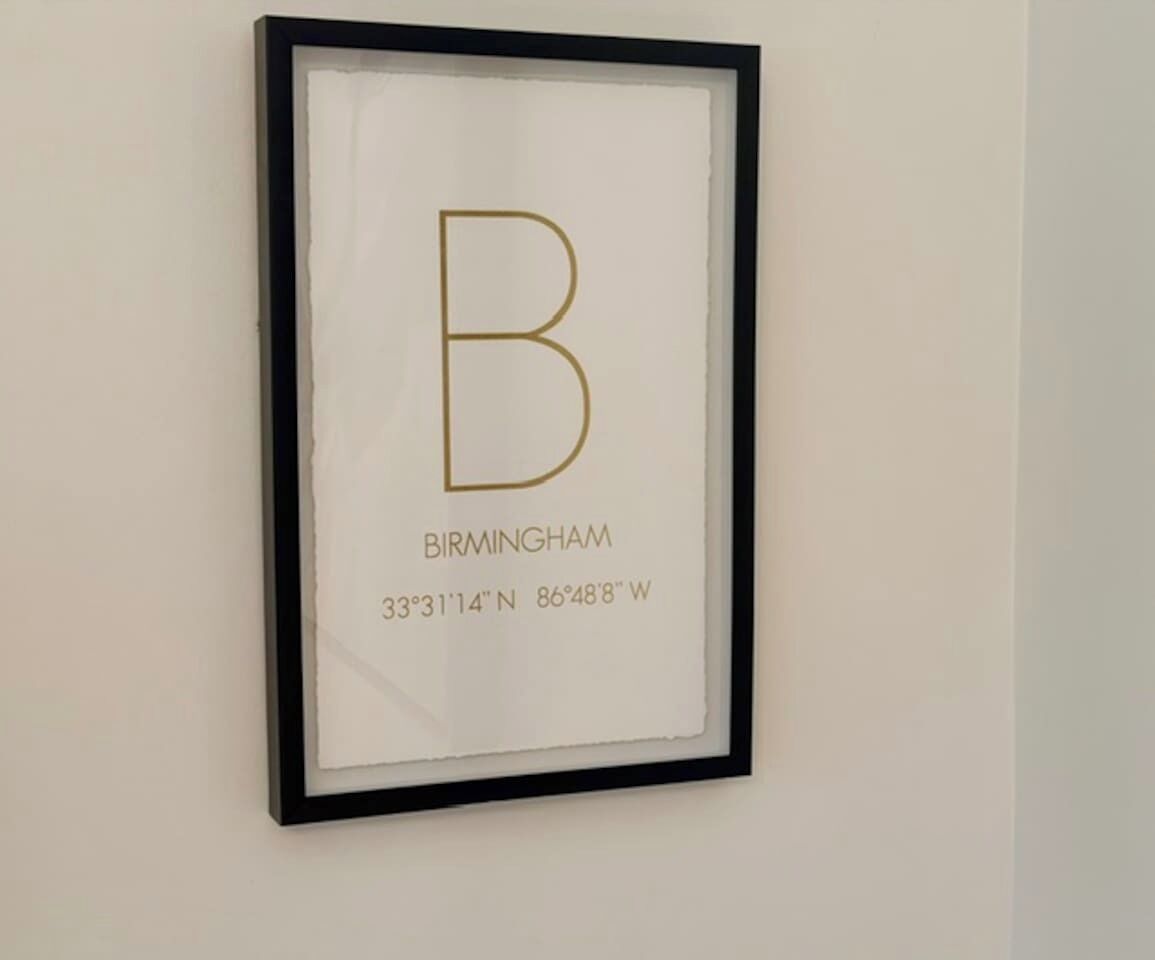 A framed artwork with a large gold letter 'B' and the text 'BIRMINGHAM 33°31'14'N 86°48'W' on a white background.