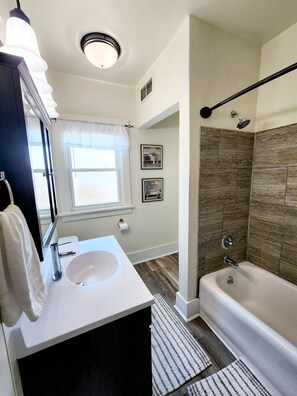 Een badkamer met een wit wastafelmeubel, een spiegel en een douche met regendouchekop, plus een raam met uitzicht.