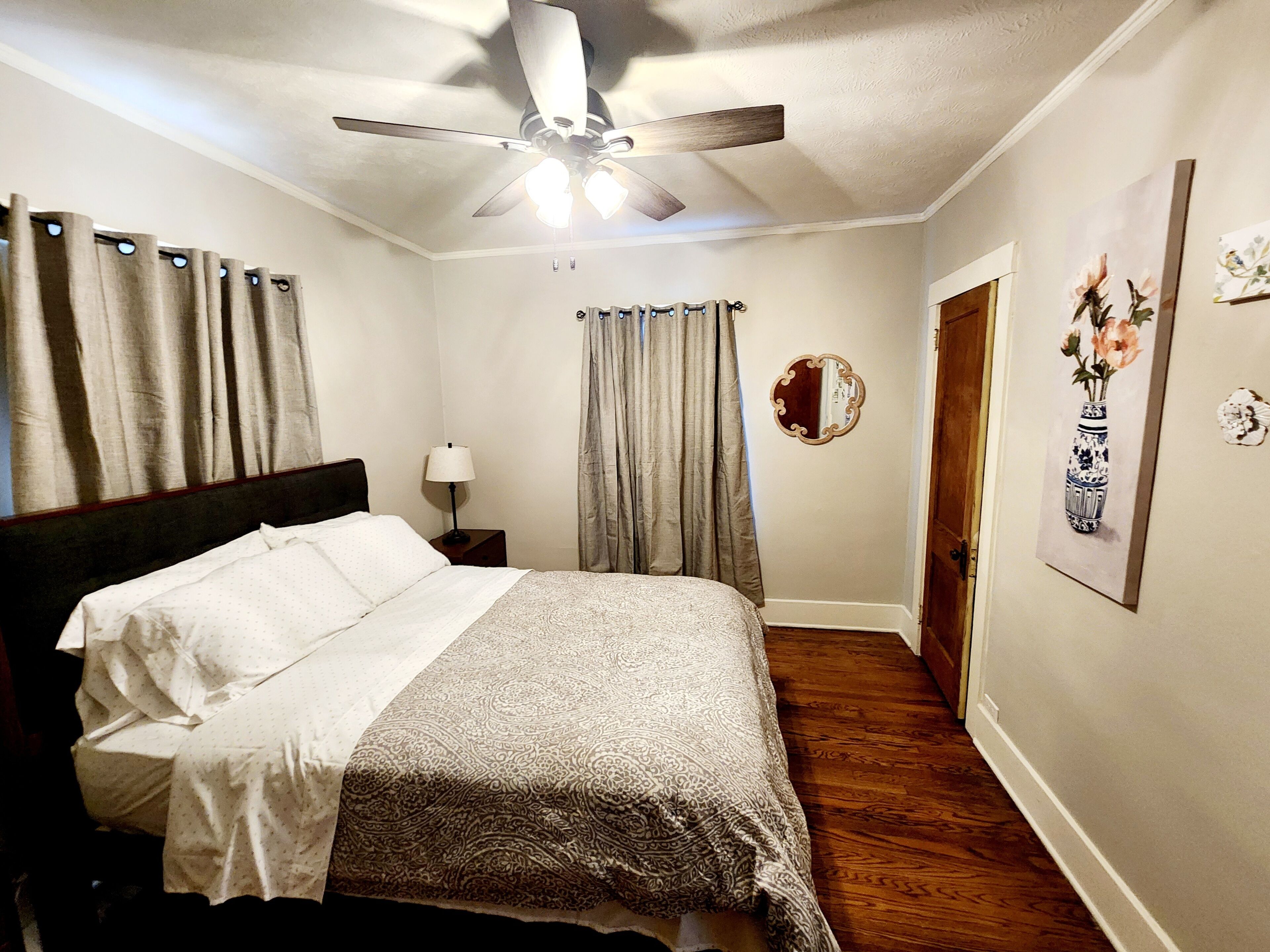A bedroom with a bed, a ceiling fan, a nightstand with a lamp, a door, and a wall with a framed picture.