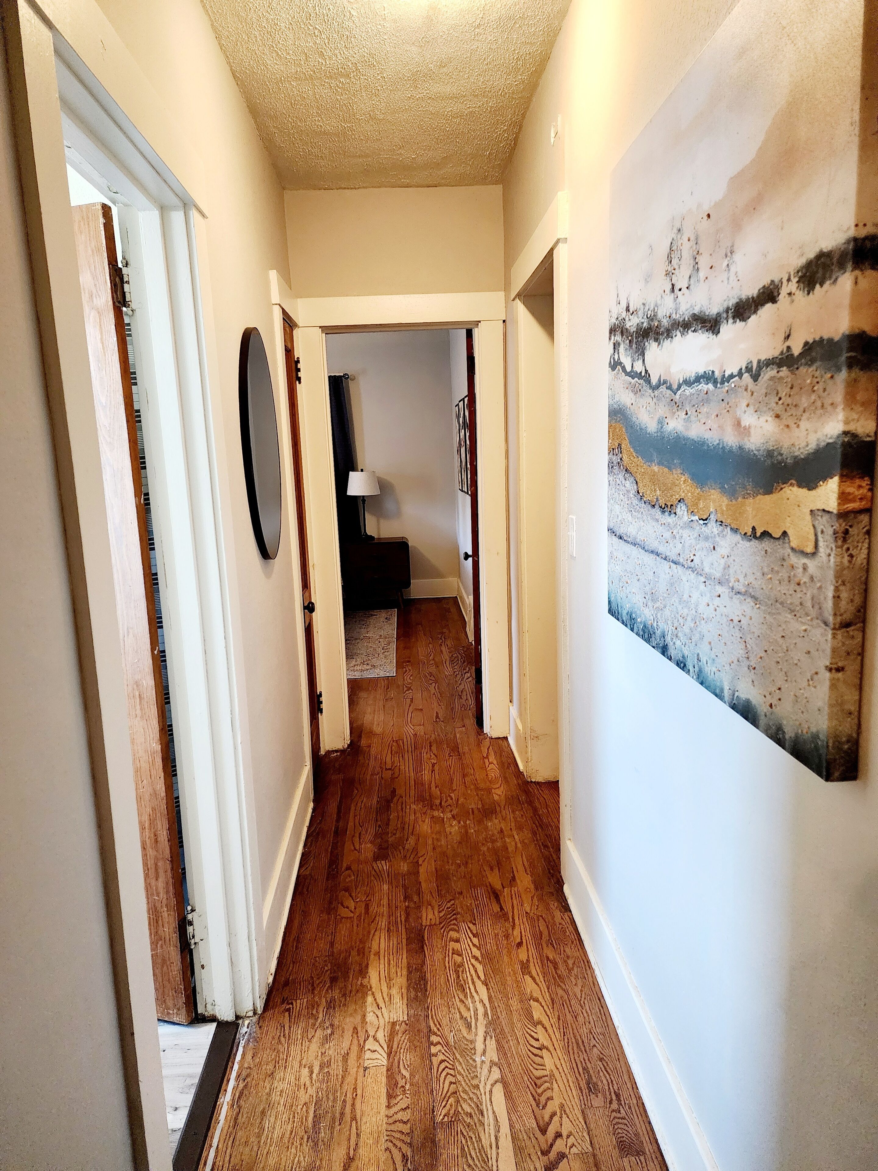 A hallway with wooden flooring, a door on the left, a wall-mounted lamp, and a painting on the right.