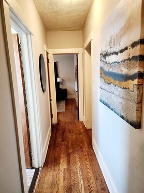 A hallway with wooden flooring, a door on the left, a wall-mounted lamp, and a painting on the right. - 2-bed house, across the street from UNMC. (Omaha)