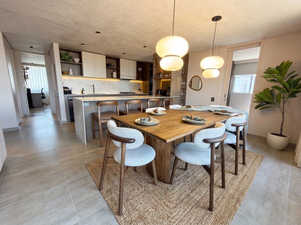 Een moderne keuken met een centraal eiland, een eettafel met stoelen, hanglampen en een potplant.