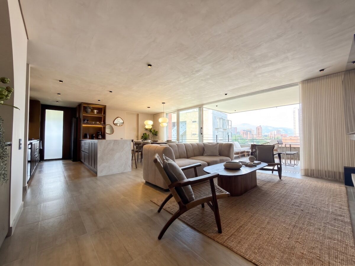 Amplia sala de estar con sofá, sillas y una mesa de centro. Se ve un área de cocina al fondo.