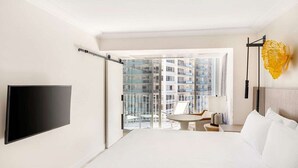 Ett hotellrum med en säng, en TV, en balkong med ett bord och stolar samt utsikt över andra byggnader.