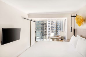 Een hotelkamer met een bed, een televisie, een balkon met een tafel en stoelen, en uitzicht op andere gebouwen.